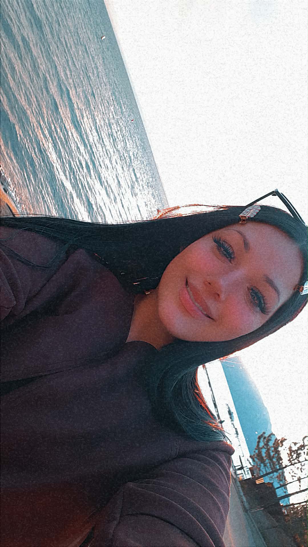 Smiling young woman with long dark hair posing for a selfie by a scenic lake.