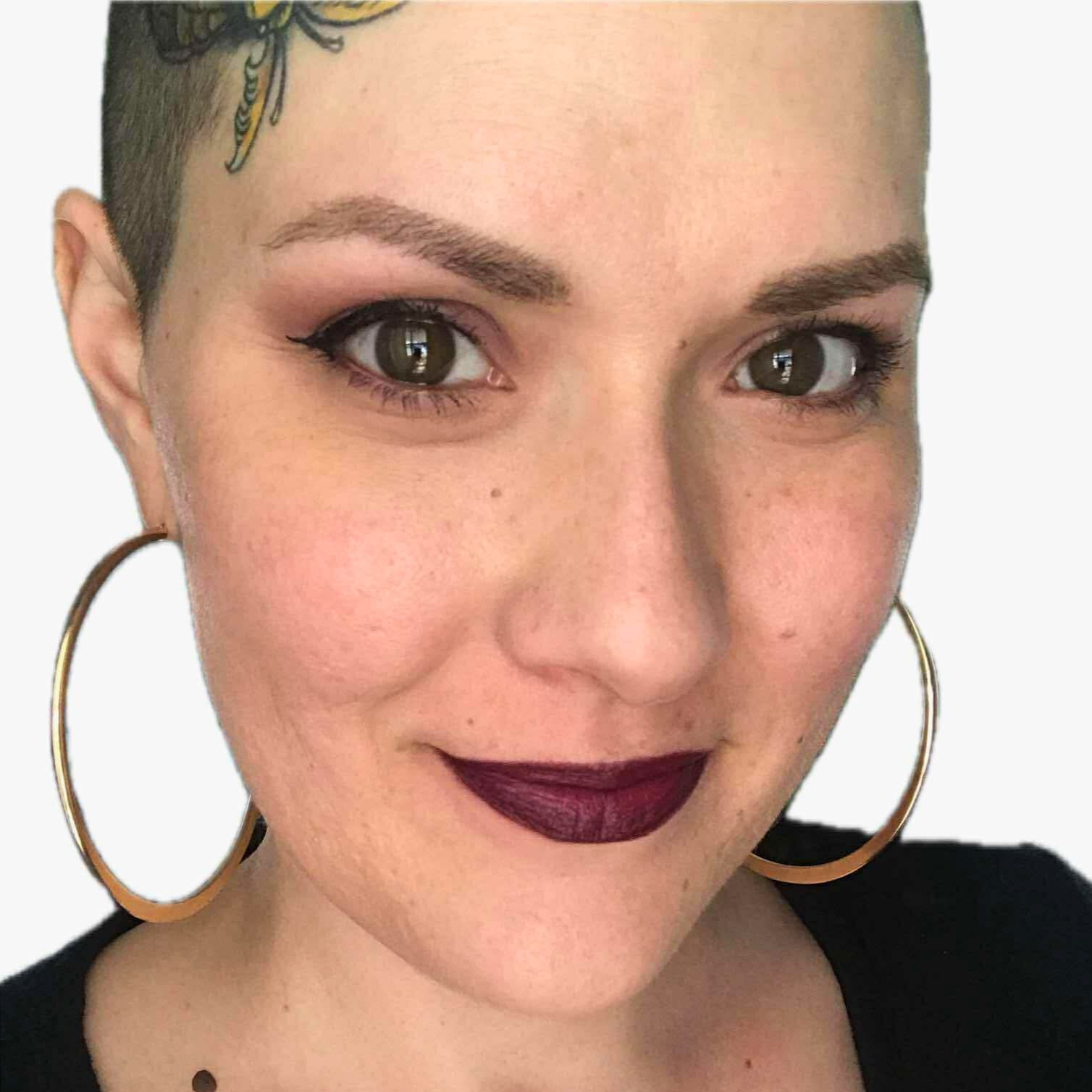 Person with a shaved head, yellow scalp tattoo, large gold hoop earrings, and burgundy lipstick.